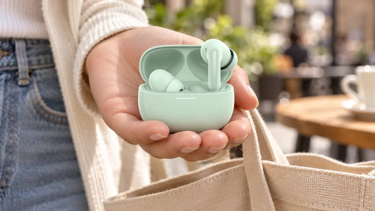 A hand holding an open case with mint wireless earbuds next to a canvas bag.